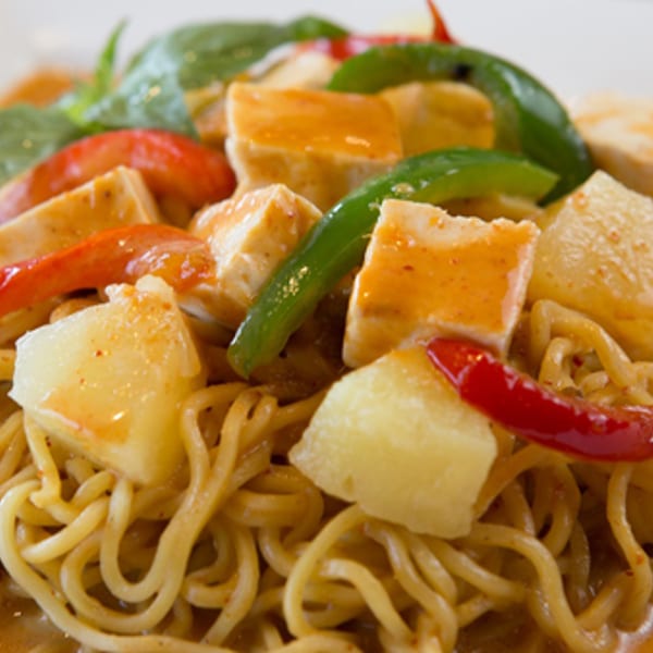 Close-up of yellow noodles in red curry sauce with tofu, pineapple, bell peppers, and basil.
